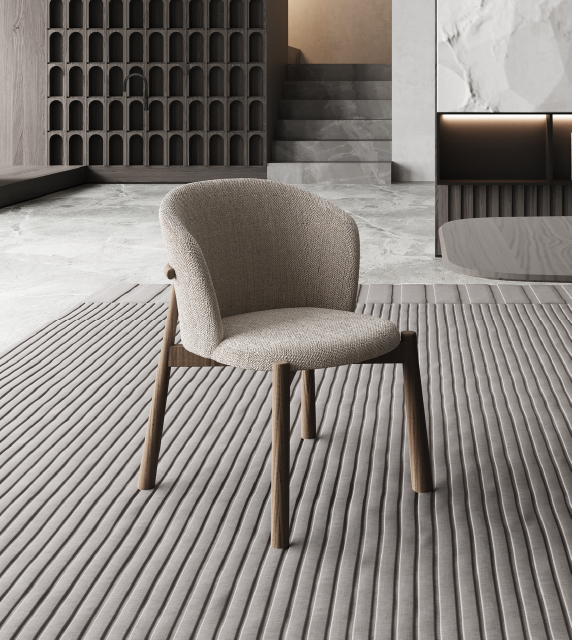 Beige upholstered lounge chair with wooden legs on a striped rug in a modern, neutral-toned living space with concrete floor and minimalist shelving in the background